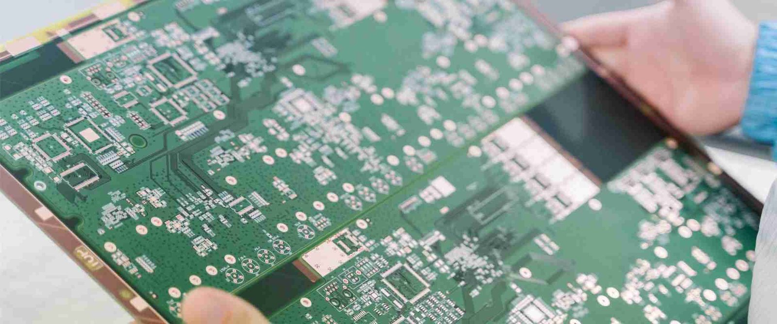 Hands holding a green bare circuit board with fine copper routing and contact pads